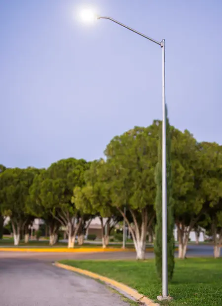 Poste metálico galvanizado de ORBE - Postes Metalicos, con un diseño de brazo simple curvo y luz LED encendida, instalado en un área verde junto a un camino.